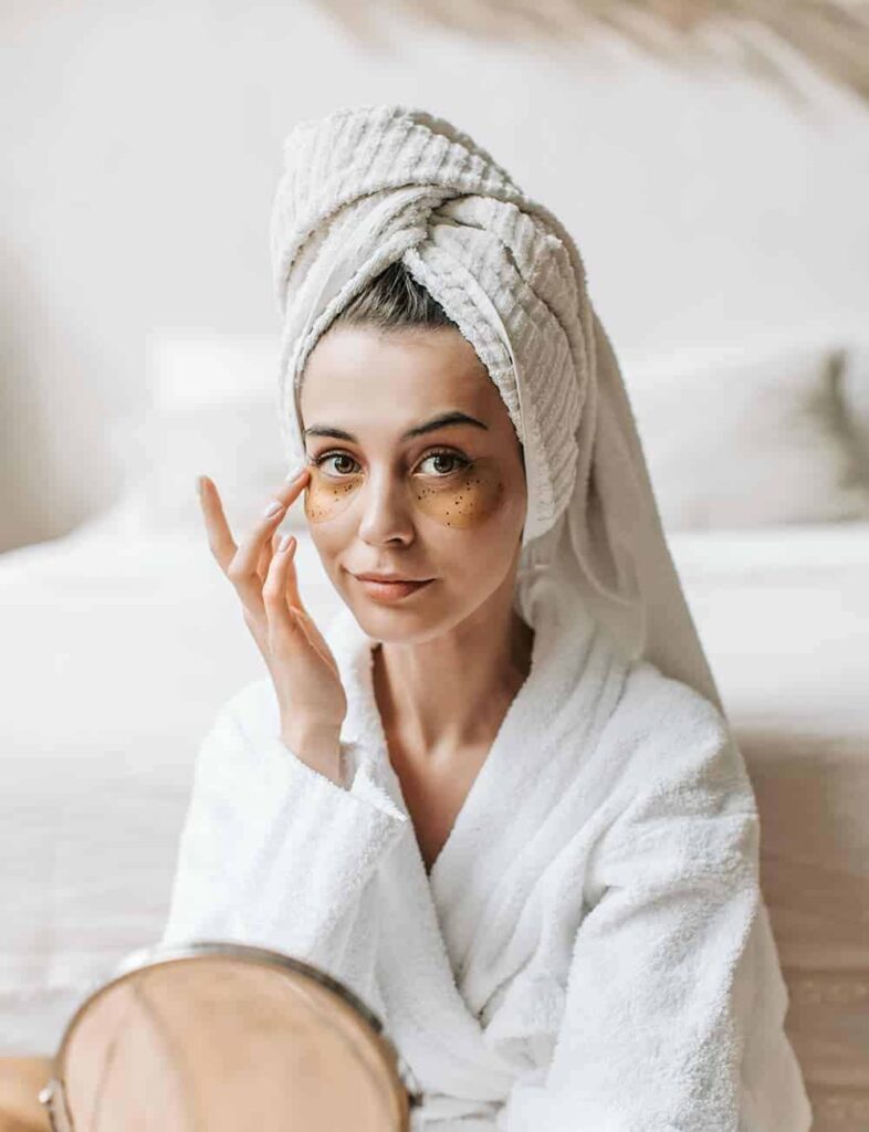 Woman in bathrobe with towel on head applying skincare cream under eyes