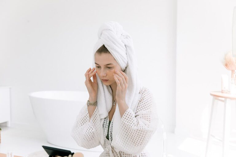 Woman in white robe and towel near bathtub touching face during skincare routine