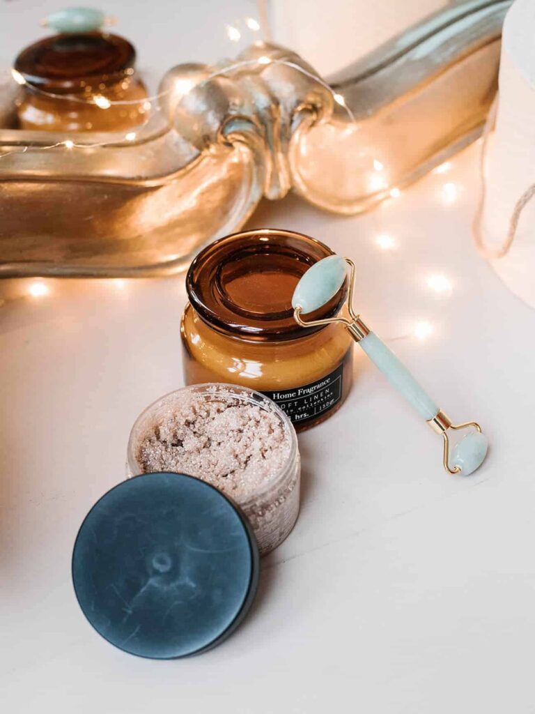 Open jar of exfoliating scrub with jade facial roller on brown glass jar, set on light surface with warm decorative lighting