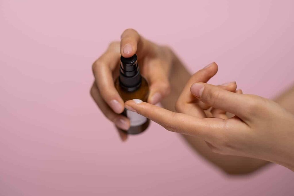 Hands dispensing skincare serum from brown pump bottle onto fingertip against pink background